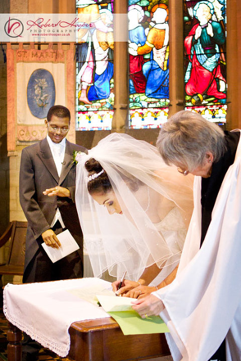 Groom examines wedding ring while bride signs register 