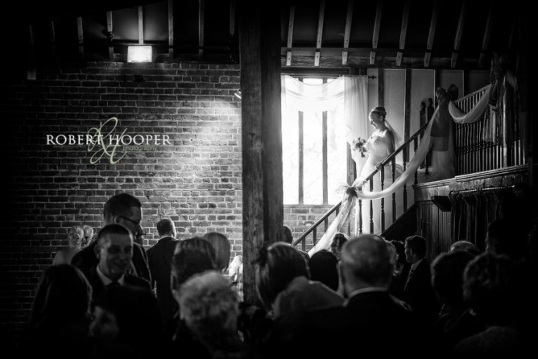 Wedding ceremony photography at Cooling Castle Barn in Rochester, Kent. Bride passes window bathed in light as she enters the wedding ceremony