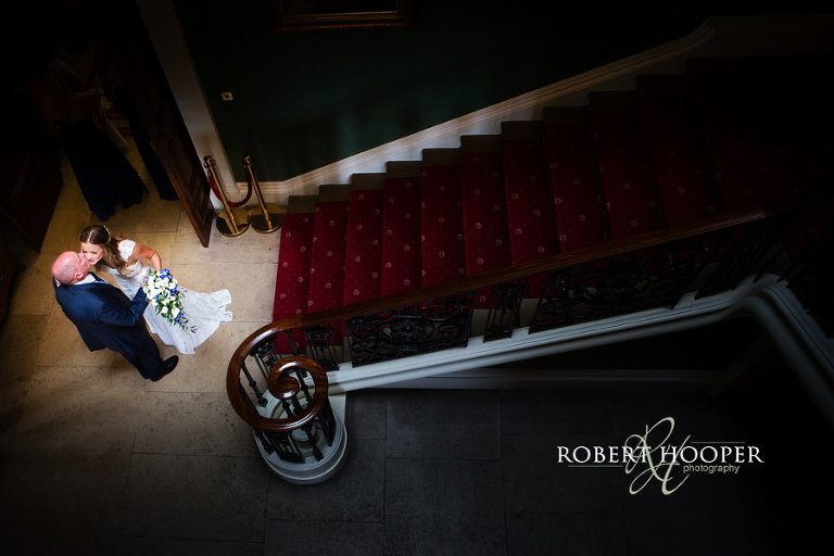Father of the bride greets his daughter with a kiss at the foot of the staircase before her wedding at Addington Palace in Surrey