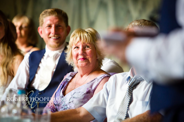 Groom's mum smiling during father of the bride's speech on the top table at the wedding breakfast in the Great Hall, Addington Palace, Surrey