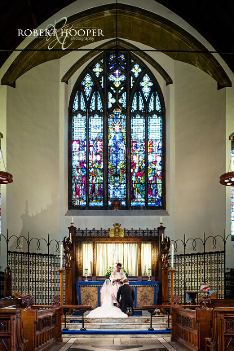 Bride and groom receiving blessing from the vicar at the alter beneath a beautiful stained glass window in St Andrew the Apostle Parish Church in Catford, London