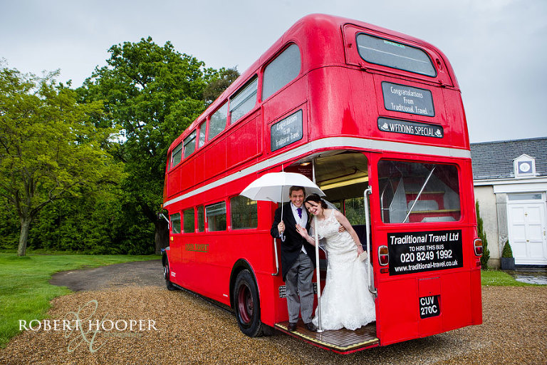 Bride and groom waving from their red London bus under a white umbrella on their rainy wedding day at Addington Palace in Surrey