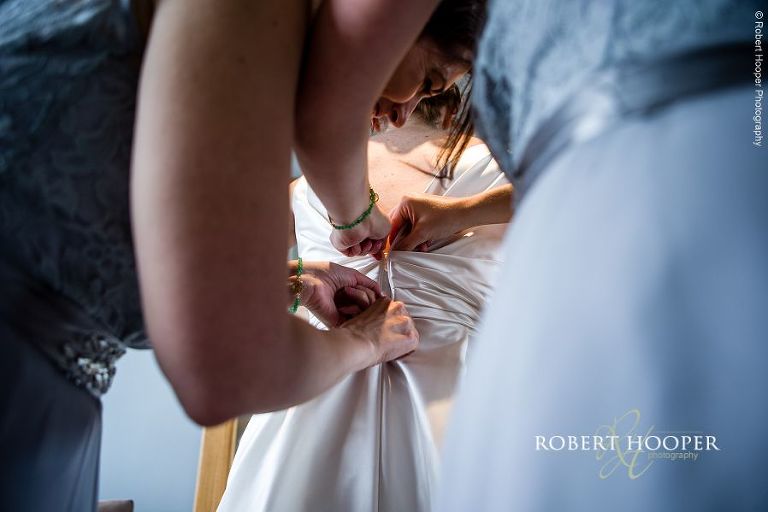 Bridesmaids helping bride put on her wedding dress on her wedding day in August at Bix Manor, Henley on Thames in Oxfordshire