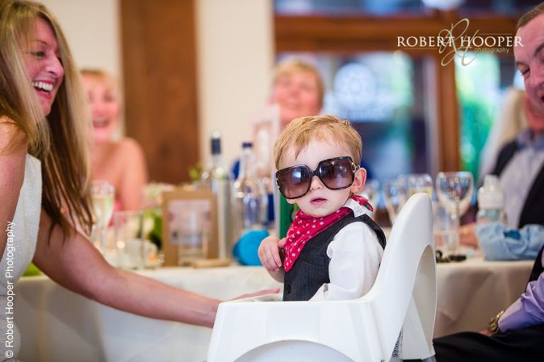 Toddler in his high chair wearing his mum's sunglasses at wedding breakfast at Coltsford Mill, Oxted in Surrey
