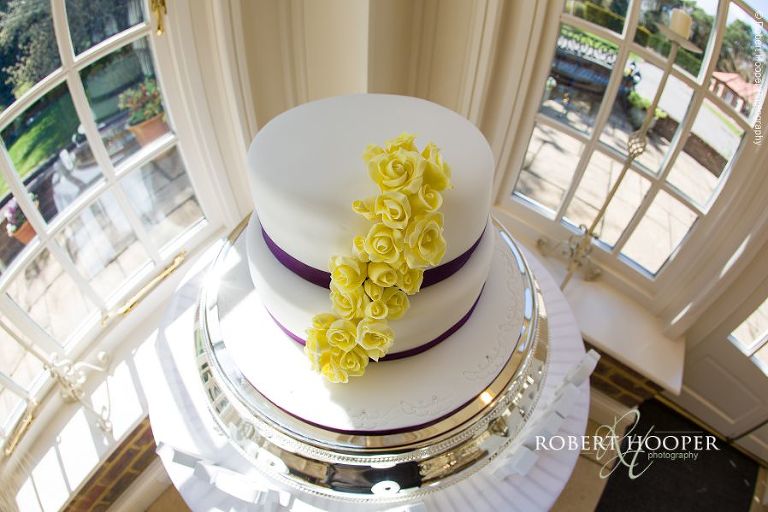 Wedding cake with flourish of yellow icing flowers down side on corner table at Oaks Farm Barn wedding venue in Surrey