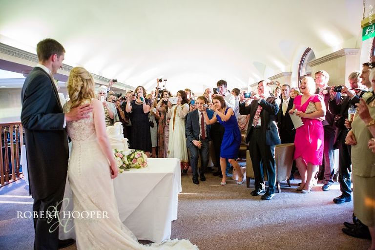 Bride and groom cut their wedding cake with all their guests watching and photographing in the Long Bar at the London Golf Club in Kent