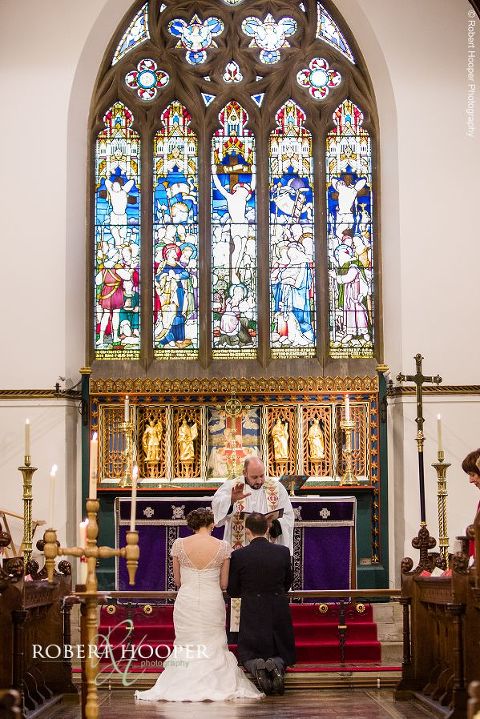 Bride and groom kneeling in front of beautiful stained glass window for blessing by vicar at St. Anne's Church in Surrey