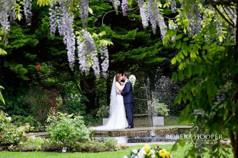Bride and groom kiss in front of pond framed with Wisteria at Oaks Farm Barn wedding venue Croydon Surrey