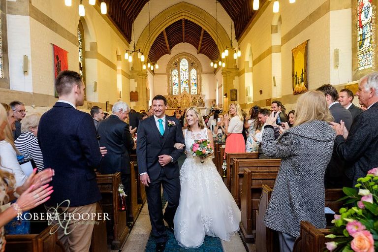 Bride and groom walk back down the isle having just wed while guests clap and take photographs at All Saints' Church Hordle Hampshire