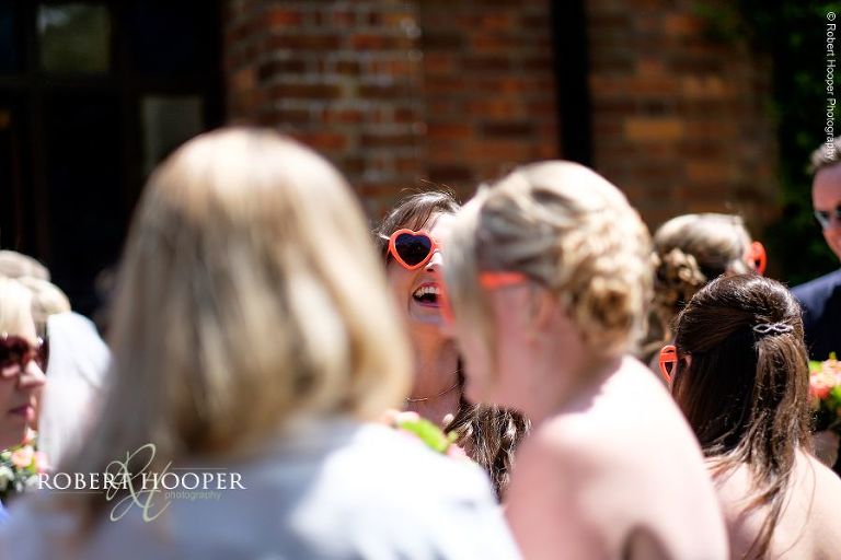 Bridesmaids wearing love heart shaped plastic orange sunglasses on sunny wedding day at All Saints' Hordle Hampshire