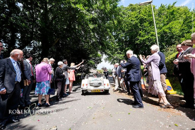 Wedding guests waving, throwing confetti and taking snaps as newlyweds leave All Saints Hordle Church for their wedding reception venue