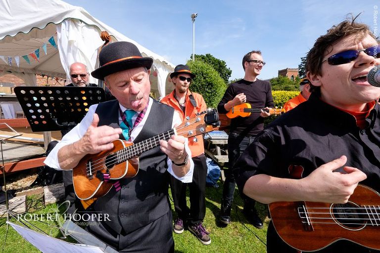 Father of the bride concentrates whilst playing ukulele with wedding band at village fayre themed wedding reception in the marquee and gardens of The Three Tuns Bransgore Dorset