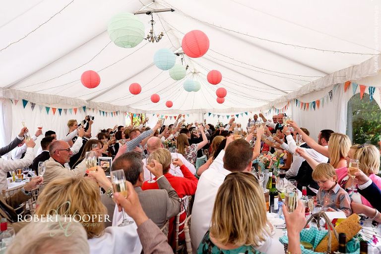 All wedding guests raising their champagne glasses in a toast to the happy couple at their wedding breakfast in the marquee at The Three Tuns Bransgore Dorset