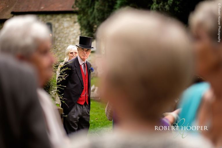 Wedding guest in top hat and tails outside St. Peter and St. Paul's church in Chaldon on summer wedding day in Surrey