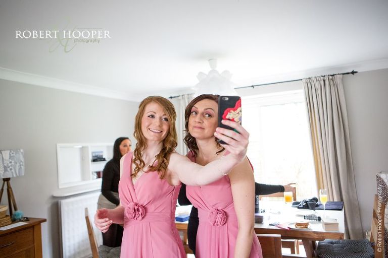 Bridesmaids in salmon pink dresses taking a selfie after their hair and make-up session for wedding day at Great Fosters Surrey