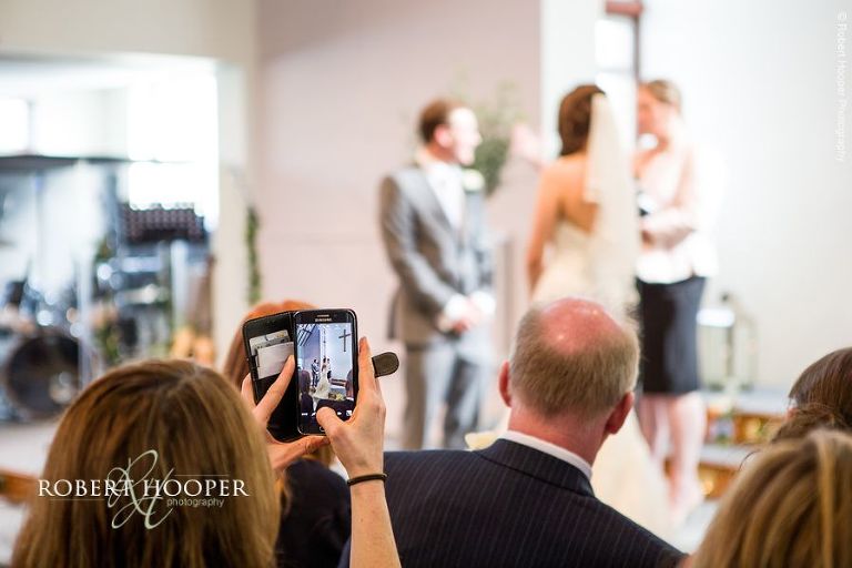Wedding guest seated in the pew taking a photo of the bride and groom with her iPhone during wedding service at Bookham Baptist Church Surrey