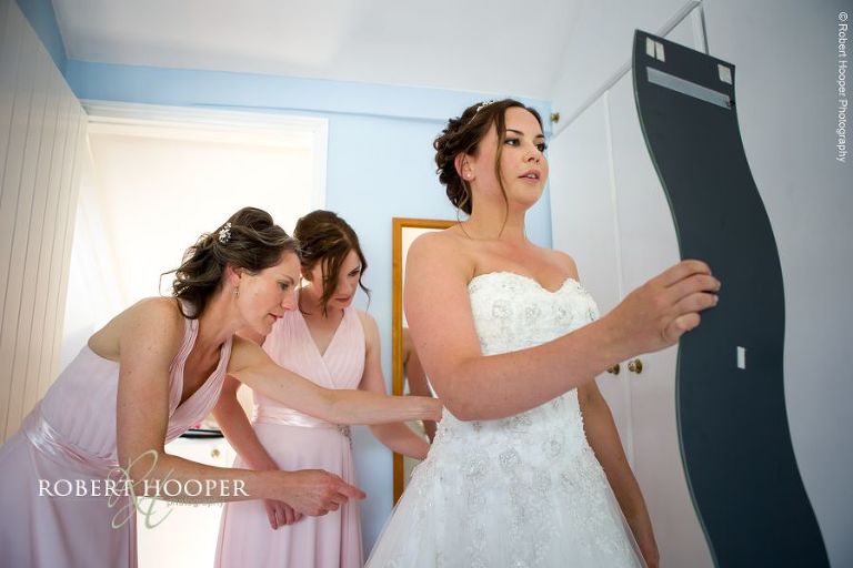Bridesmaids helping bride tie up back of wedding dress before ceremony in the barn at The Crown Inn Pishill Oxfordshire