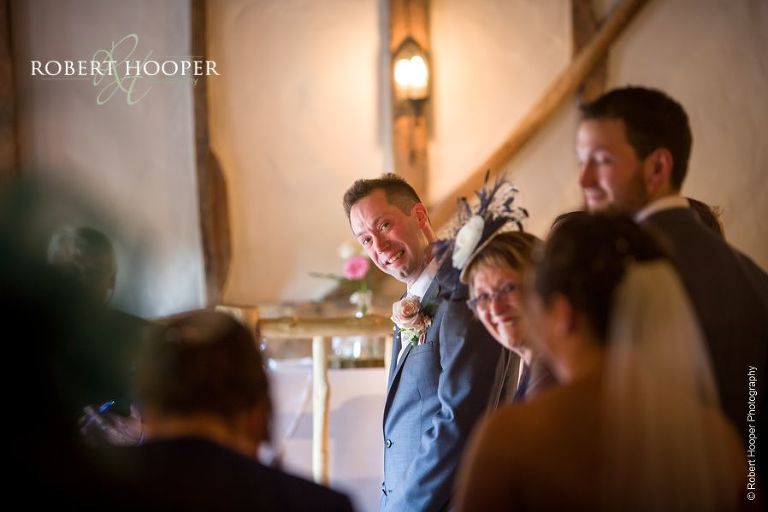 Groom looks down the isle with a smile for his bride-to-be approaching for their wedding ceremony in the barn at The Crown Inn Pishill Oxfordshire